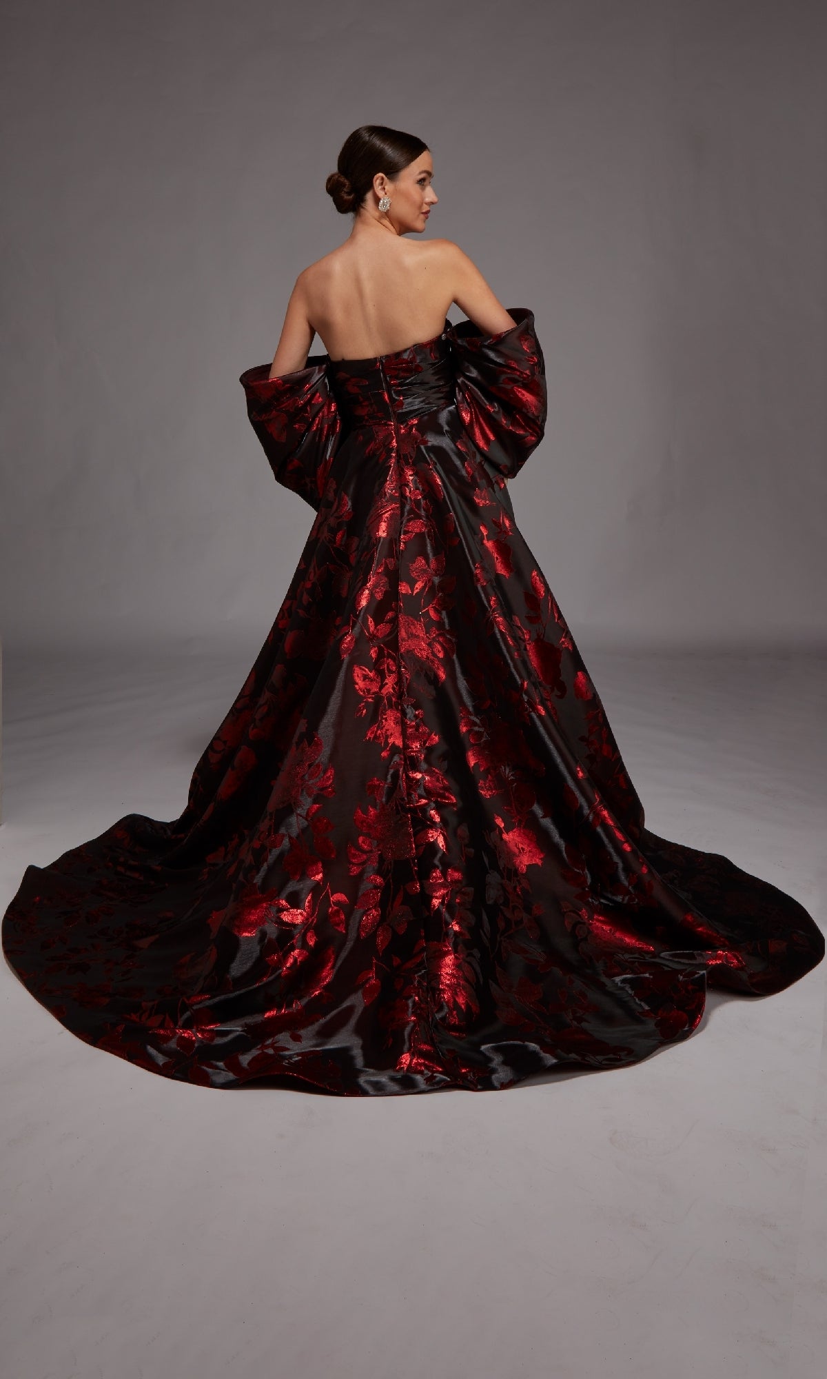 Woman wearing the Alyce 27862 long formal dress—a stunning strapless black floral jacquard gown with red patterns and a long train—stands with her back to the camera against a plain background.